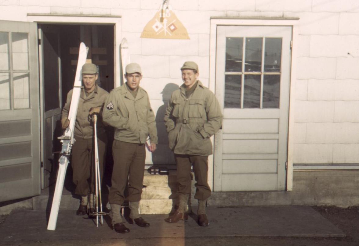 three Members of the Tenth Mountain Division, 110th Signal Corps Company, stand outside the door of their barracks at Camp Hale, Colorado. All are in uniform and one is carrying skis and poles